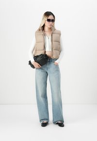 Kvinne med beige polstret vest, hvit kort topp, vide jeans, svarte leiligheter, store solbriller, som holder på en svart clutch-veske.