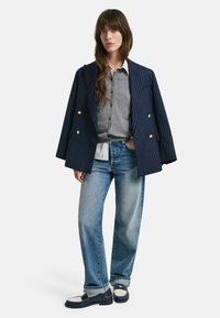 Blazer oversize a righe blu navy, maglione grigio a bottoni, jeans azzurri chiari e mocassini blu navy con dettaglio bianco. Look casual e stratificato.
