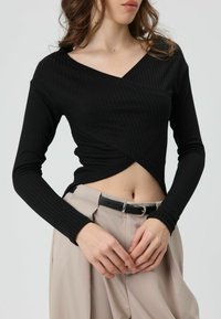 Zwart geribd topje met lange mouwen, een V-hals en een asymmetrisch wikkelontwerp, gecombineerd met lichtbeige broek met hoge taille en een zwarte riem.