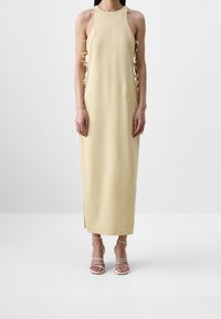 Femme portant une longue robe beige sans manches avec des détails lacés sur les côtés et des sandales blanches à talons hauts et brides, debout sur un sol blanc.