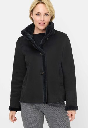 Frau trägt eine schwarze Jacke mit Pelzbesatz und Knopfverschluss sowie schwarz-weiß karierte Hose, steht vor weißem Hintergrund.