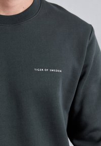 Hanorac de culoare verde închis, fabricat dintr-un material moale, cu guler rotund și textul "TIGER OF SWEDEN" în alb pe piept.