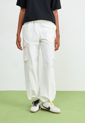 Pantalon cargo blanc avec taille élastique, poches latérales et coupe décontractée, associé à des baskets noir et blanc sur un sol vert.