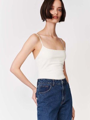 Lühikeste tumedate juustega naine, kes kannab valget spageti rihmadega toppi ja kõrge vöökohaga siniseid teksapükse, seisab ühe käega taskus tavalise tausta ees.