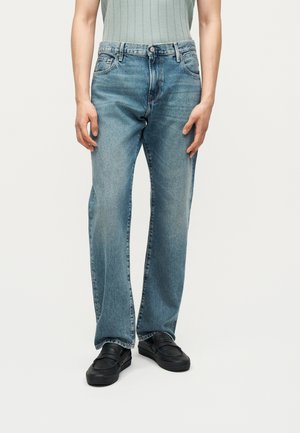 Osoba nosząca jasnoniebieskie jeansy o prostym kroju, czarne wsuwane buty i włożony do środka prążkowany top bez rękawów, stojąca na jednolitym tle.