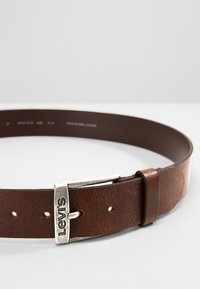 Cinturón de piel marrón con una hebilla de metal en tono plateado grabada con "Levi's". El cinturón tiene agujeros para ajuste y una textura suave.