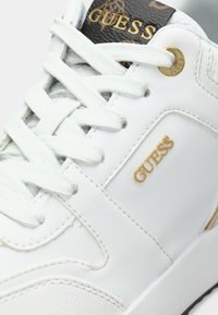 Sneaker sintetico bianco con lacci piatti, accenti dorati e una patches con logo nera. Presenta una superficie liscia e una forma della punta arrotondata.