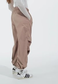 Beige, loose-fitting pants with an elastic waist and side pockets, paired with white sneakers featuring black stripes. Smooth texture.