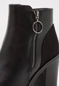 Stivaletto alla caviglia nero con tacco realizzato in pelle liscia, dotato di una zip laterale con un tirante circolare e un accentuato tessuto testurizzato. Design semplice, profilo elegante.