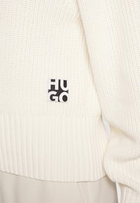 Primo piano di un maglione a coste color crema con un piccolo patch del marchio "HUGO" in bianco e nero vicino alla vita.