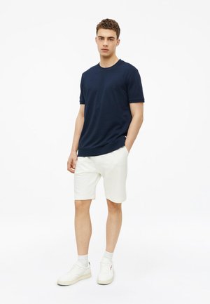 Junger Mann trägt ein marineblaues kurzärmeliges Hemd, weiße Shorts, weiße Socken und weiße Turnschuhe, steht mit einer Hand in der Tasche vor weißem Hintergrund.