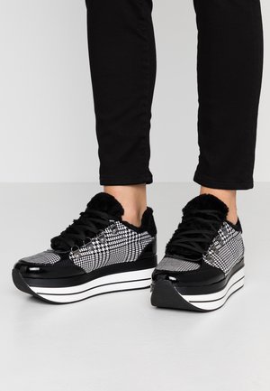Czarno-białe sneakersy na platformie w kratę pepitkę z futrzaną wyściółką i czarnymi sznurowadłami, noszone z czarnymi spodniami na neutralnym tle podłogi.