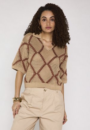 Donna con capelli ricci che indossa un top lavorato a maglia beige e marrone con motivo a losanghe e pantaloni beige chiaro a vita alta.