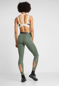 Sportbeha met een kleurenschema van roze en olijfgroen; groene legging met een ontwerp van mesh uitsnijdingen en een zwarte tailleband met de tekst "NIKE PRO".