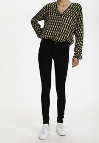 Femme portant un chemisier à manches longues à motifs, un pantalon skinny noir et des baskets blanches, se tenant avec une main sur la hanche contre un fond blanc.