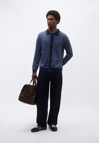 Μπλε πλεκτή ζακέτα με σχέδια, σκοτεινό γιακά και κουμπιά, συνδυασμένη με σκούρο παντελόνι με φαρδιά μπατζάκια και μαύρα αθλητικά παπούτσια, κρατώντας καφέ δερμάτινη τσάντα.