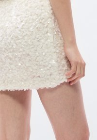 Φόρεμα σε ανοιχτό κρεμ χρώμα με υφή, καλυμμένο με μικρές παγιέτες. Μήκος πάνω από το γόνατο, εφαρμοστή γραμμή και σκάλιστη τελείωμα.