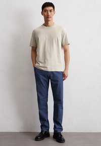 Cremfarbendes Baumwoll-T-Shirt mit kurzen Ärmeln und kleinem Logo, kombiniert mit dunkelblauen Jeans und schwarzen Schnürschuhen.