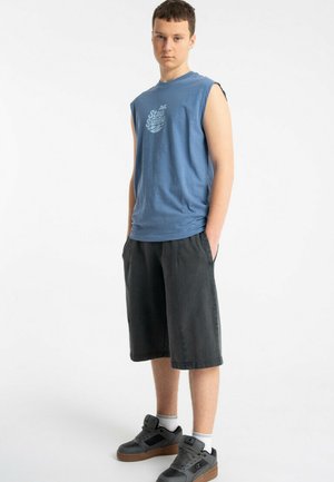 Młody mężczyzna w niebieskiej koszulce bez rękawów z napisem "Stay Sunny", czarnych spodenkach za kolano, białych skarpetkach i czarnych tenisówkach, stoi z rękami w kieszeniach.