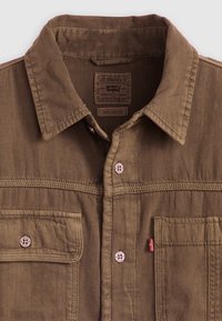 Camisa de trabajo marrón hecha de tela con acabado texturizado. Presenta un cuello, bolsillos frontales y herrajes de marca. Diseño de corte relajado.