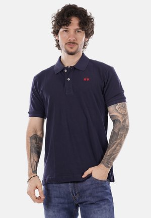 Uomo con capelli ricci che indossa una polo blu navy e jeans blu, che mostra tatuaggi dettagliati su entrambi gli avambracci, posando contro uno sfondo bianco.