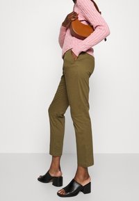 Oliwkowe dopasowane spodnie z przednimi kieszeniami, zestawione z jasnoróżowym swetrem w warkocze i czarnymi mule na obcasie z odkrytymi palcami. Brązowa torebka na ramię.