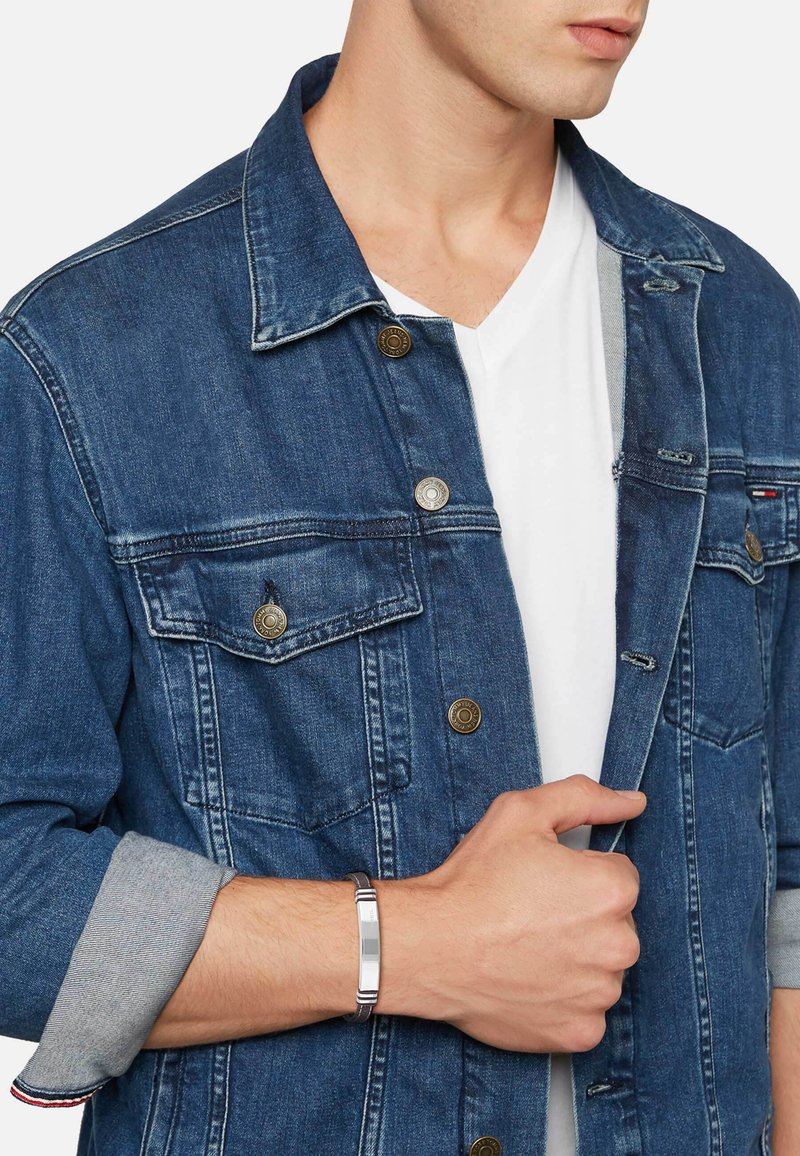 Man som bär en blå denimjacka över en vit t-shirt, håller jackan öppen med ena handen, bär ett silverarmband på handleden.