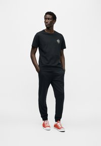 Μαύρο βαμβακερό t-shirt με μικρό γραφικό φυτού, σε συνδυασμό με μαύρο παντελόνι φόρμας και κόκκινα αθλητικά με λευκά κορδόνια. Απλό και άνετο fit.