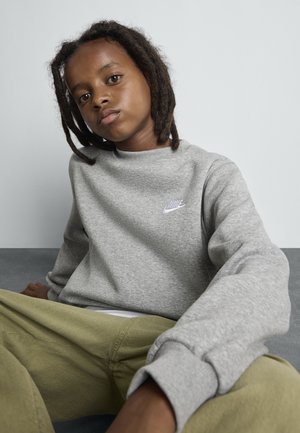 Mladá osoba s dredmi, oblečená v šedé mikině Nike a olivových nohaviciach, sedí uvoľnene proti jednolitému svetlému pozadiu.