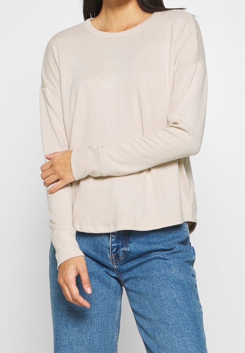 Personne portant un t-shirt beige à manches longues et un jean bleu, avec un bras croisé et la main reposant sur le coude opposé.