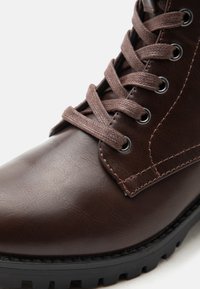 Brauner Lederstiefel mit dunkelbraunen Schnürsenkeln, die durch Metallösen gezogen sind, und einer dicken schwarzen Sohle mit tiefem Profil.