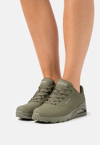 Olivgrüne Skechers-Sneaker mit strukturiertem Obermaterial, Schnürdesign, perforierten Details und sichtbarer Luftpolsterung in der Sohle.