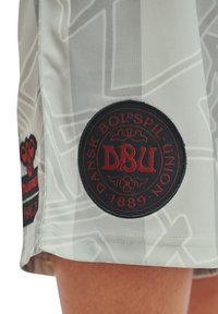 Röd och svart DBU Danska Fotbollsförbundet 1889-patch på grå mönstrade sportshorts som bärs av en person.