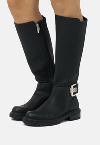 Botas de cuero negras hasta la rodilla, con cierre lateral, suela gruesa y una hebilla decorativa plateada en la pantorrilla.