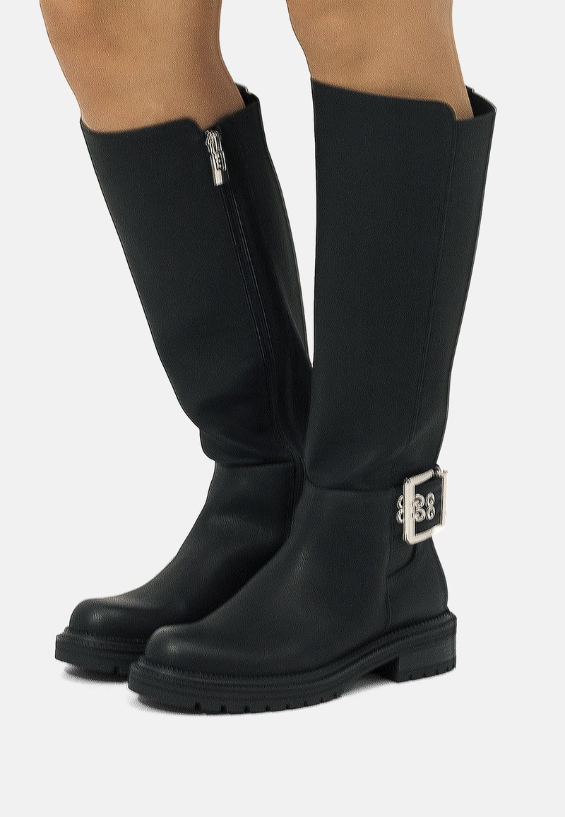 Botas de cuero negras hasta la rodilla, con cierre lateral, suela gruesa y una hebilla decorativa plateada en la pantorrilla.