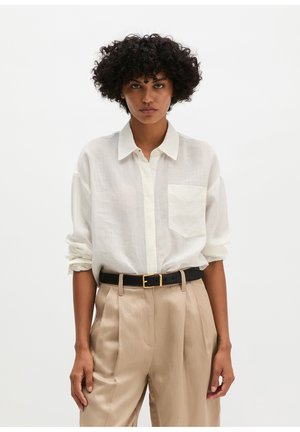 Person mit lockigem Haar, die ein weißes Hemd mit Knöpfen und hochgekrempelten Ärmeln trägt, beige Hose mit hoher Taille und einen schwarzen Gürtel mit goldener Schnalle.