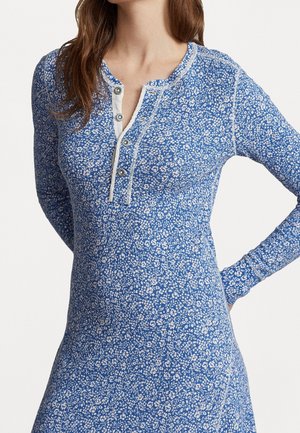 Femme portant une robe bleue à manches longues avec un imprimé floral blanc et une patte boutonnée à l'encolure, un bras plié à la taille.