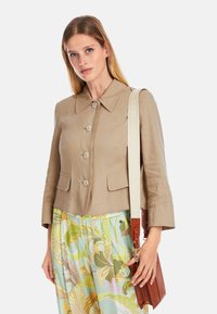 Beige cropped jacket with a wide collar, four buttons, and two front pockets. Paired with colorful floral-patterned pants and a brown bag.