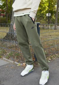 Olivgröna Nike-träningsbyxor med sidan fickor och logotyp. Kombineras med vita och gröna Nike-sneakers med nät- och gummisulor.