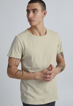 Camiseta corta de manga corta color beige claro, fabricada en tela texturizada, con un escote redondo y puños de manga enrollados.