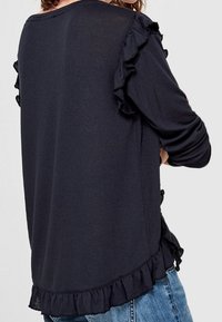 Svart långärmad topp med volangdetaljer längs axlarna och nederkanten, tillverkad av mjukt, lätt material med en avslappnad passform.