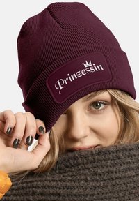 Maroon knitted beanie featuring a rectangular patch with "Prinzessin" in white script, accented by a crown. Soft texture, ribbed design.