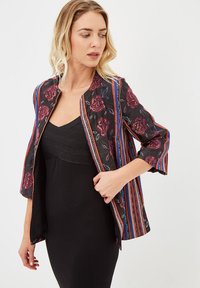 Mujer rubia ajustando una chaqueta abierta de flores y rayas sobre un vestido negro ajustado frente a un fondo liso.