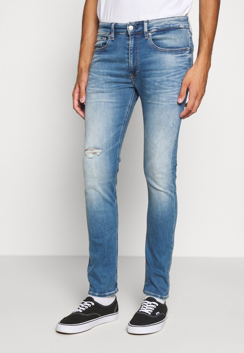 Vaqueros de mezclilla azul claro con un corte ajustado, que incluyen una rodilla desgastada, un lavado descolorido y diseño clásico de cinco bolsillos. Combinados con zapatillas negras.