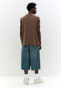 Top senza maniche marrone abbinato a pantaloni corti di jeans blu larghi, con tasche posteriori e orli risvoltati. Le calzature sono sneakers chiare con calzini bianchi.