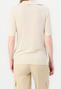 Maglione polo beige a maniche corte con colletto, orlo a costine e texture liscia, abbinato a pantaloni cargo color cammello. Il design presenta dettagli di cucitura sottili.