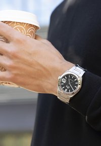 Reloj de acero inoxidable plateado con un dial negro, correa metálica y pantalla de fecha, llevado en una mano que sostiene una taza de café marrón con tapa blanca.