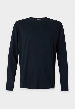 Fiatal férfi áll, tengerészkék hosszú ujjú inget, kék farmert, sportcipőt visel, és egy kockás inget kötött az oldalára.