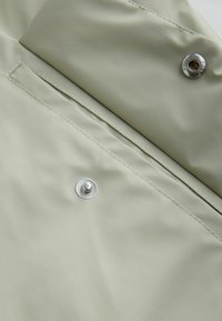 Gaiši pelēks ūdensnecaurlaidīgs audums ar gludu tekstūru, kas aprīkots ar sudraba sprādzīti un kontrastējošu šuvi. Dizaina tuvplāns.