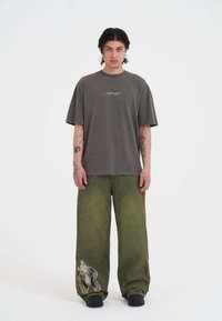Grå oversized t-shirt med et hvidt grafisk motiv på brystet, parret med løse olivengrønne bukser med et vinget grafisk design.